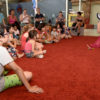 Crianças sentadas sobre tapete, assistindo a uma apresentação de contação de histórias