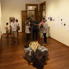 Sala de exposição, com quadros nas paredes, objetos como sacolas e pacotes no centro, e pessoas conferindo