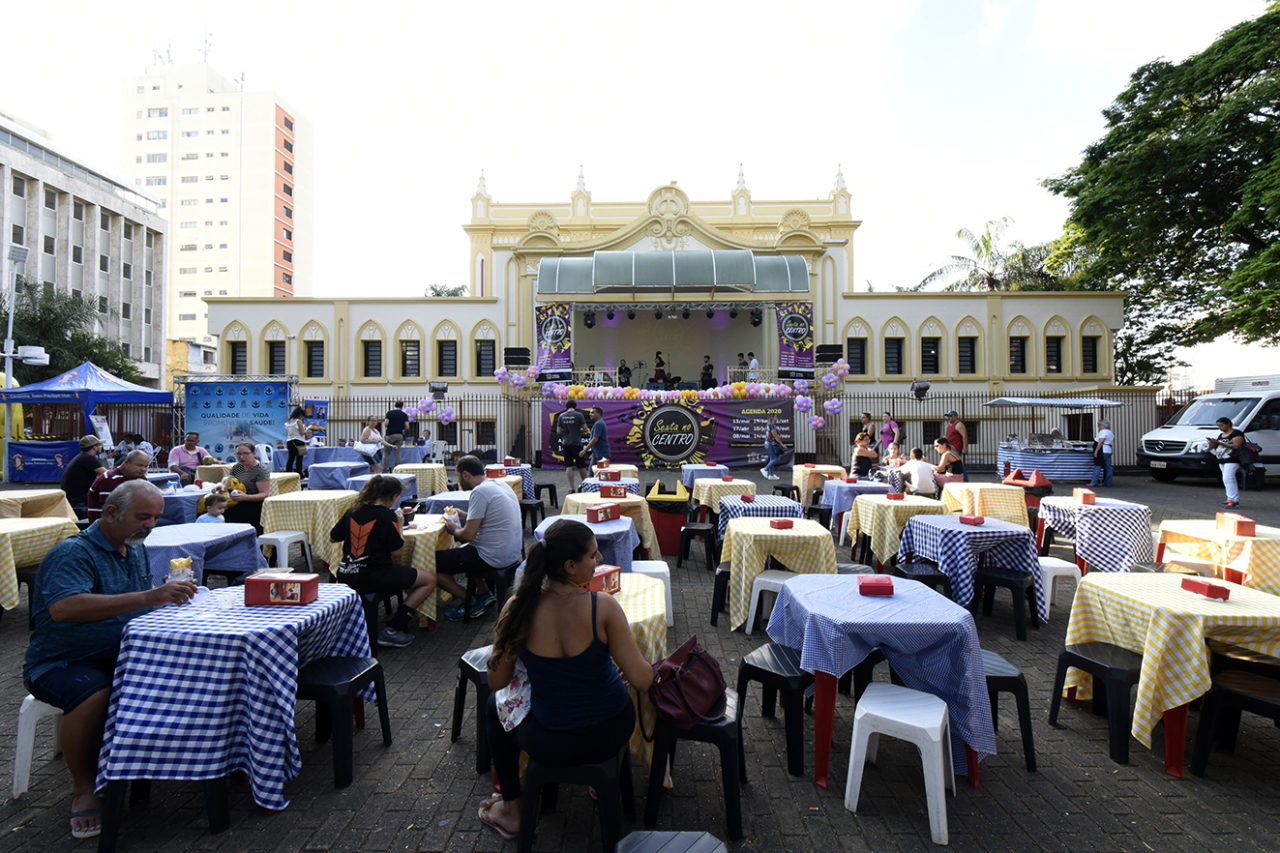 Praça com coreto ao fundo e mesas, com pessoas sentadas comendo