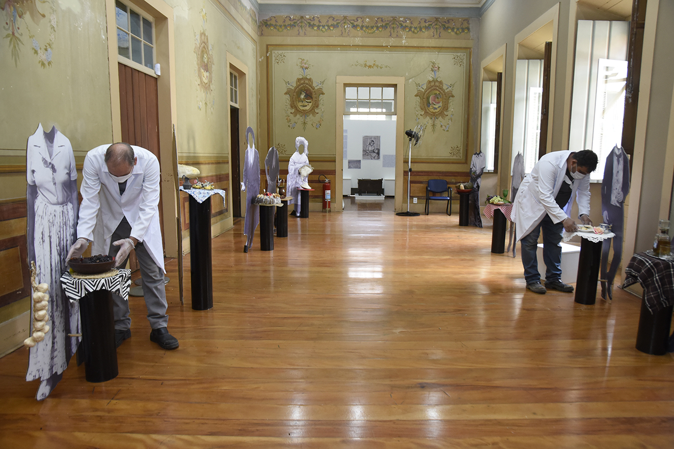 Dois homens vestindo jalecos, luvas e máscaras, montando exposição em sala com personagens de papelão aos lados