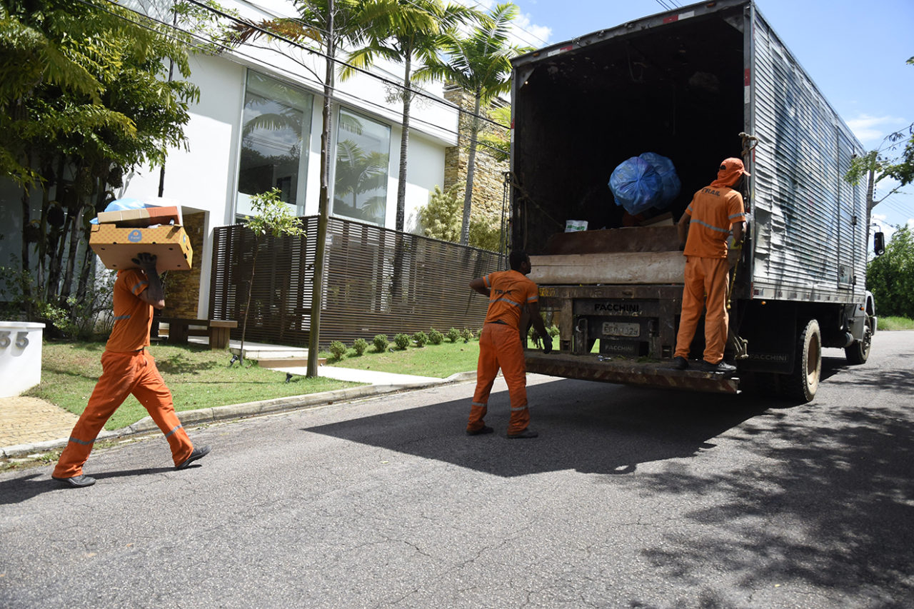 Coletores de lixo correm atrás de caminhão, um deles segurando caixas de papelão, um arremessando um saco para dentro do caminhão e outro agarrado à carreta