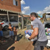 Homem retirar cestas básicas de porta-malas de carro para entregar a uma mulher e três crianças em frente auma casa de tijolos com carro na garagem