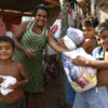 Foto posada de mulher com três meninos, segurando cestas básicas, em frente a uma casa