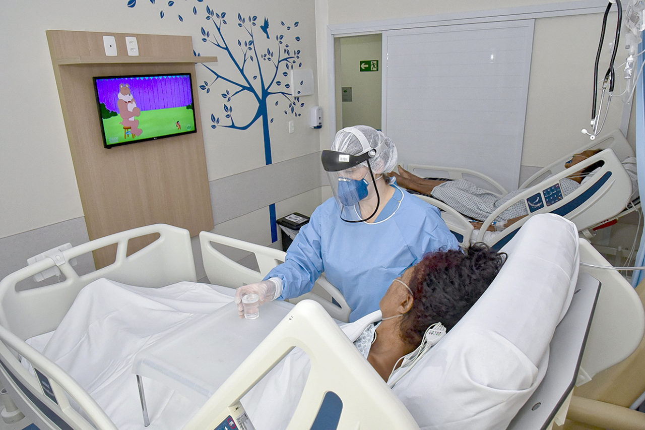 Quarto de hospital com duas pacientes e profissional da saúde uniformizada atendendo uma delas, com tela de TV ao fundo passando desenho animado