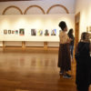 Sala de exposição de quadros, com três mulheres analisando as obras na parede da direita