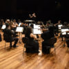 Músicos de orquestra em palco, com mulher regendo em pé e teatro em fundo escuro