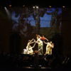 Palco de teatro no escuro, com músicos de orquestra se apresentando, enquanto atores sob feixe de luz reproduzem imagem projetada sobre a parede de fundo