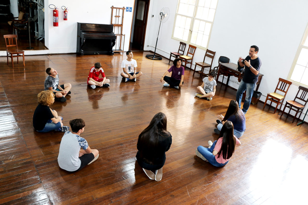 Tablado de madeira, com pessoas em círculo em atividade de aula