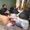 Mulher com avental e máscara abaixada, conversando com dois homens deitados sobre cobertores e caixas sobre uma calçada, apoiados em parede de azulejos