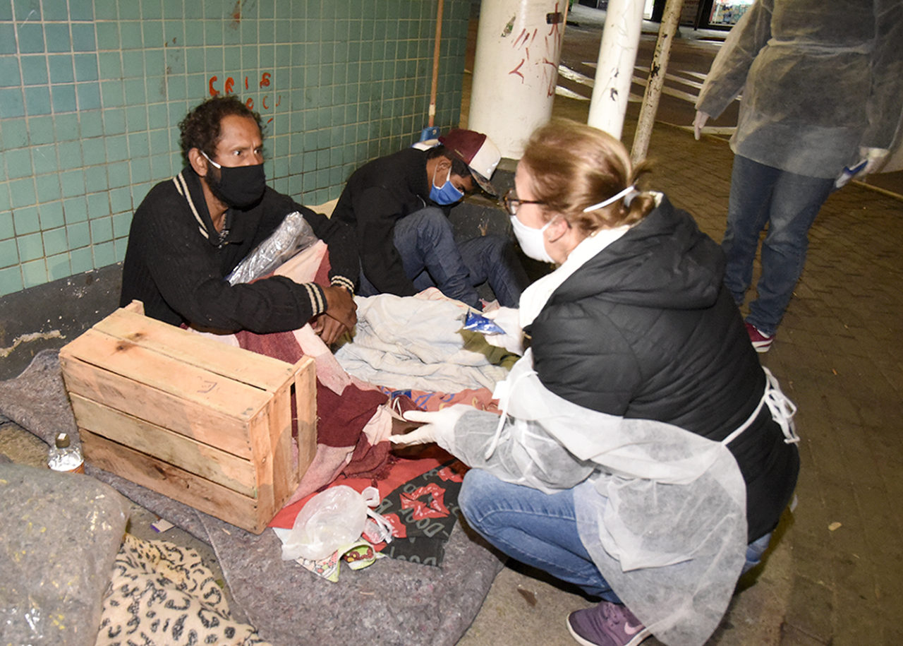 Mulher com avental e máscara abaixada, conversando com dois homens deitados sobre cobertores e caixas sobre uma calçada, apoiados em parede de azulejos