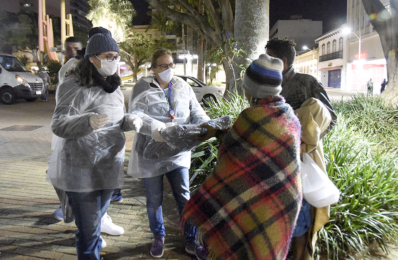 Duas pessoas com aventais e máscaras entregam pacotes a pessoas enroladas em cobertores, em uma praça, à noite