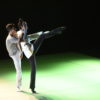 Um casal de bailarinos faz performance sobre palco, um em frente ao outro, ambos com as pernas esquerdas levantadas