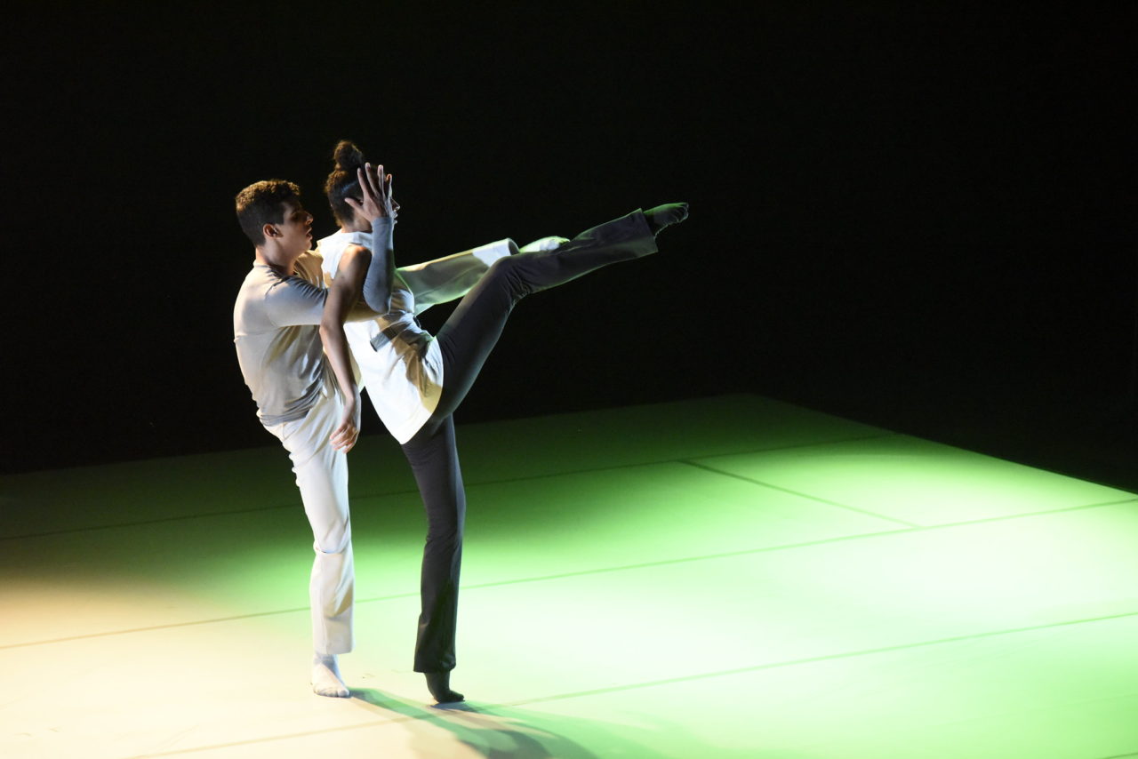 Um casal de bailarinos faz performance sobre palco, um em frente ao outro, ambos com as pernas esquerdas levantadas