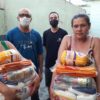 Duas mulheres à frente segurando cestas básicas com alimentos, com dois homens atrás usando máscaras