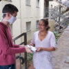 Homem usando máscara entrega pacotes para mulher, com escadaria de prédios ao fundo
