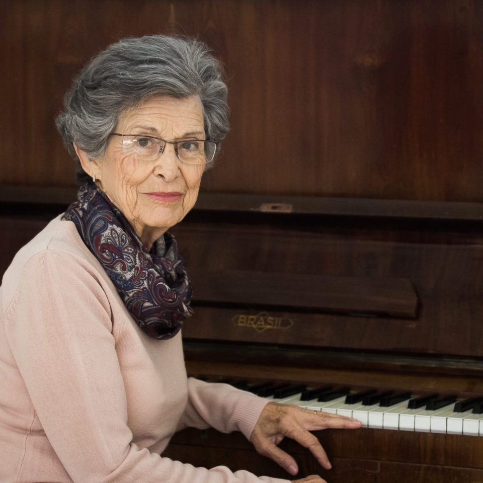 Mulher de cabelo grisalho sentada, apoiando braço esquerdo em teclas de piano