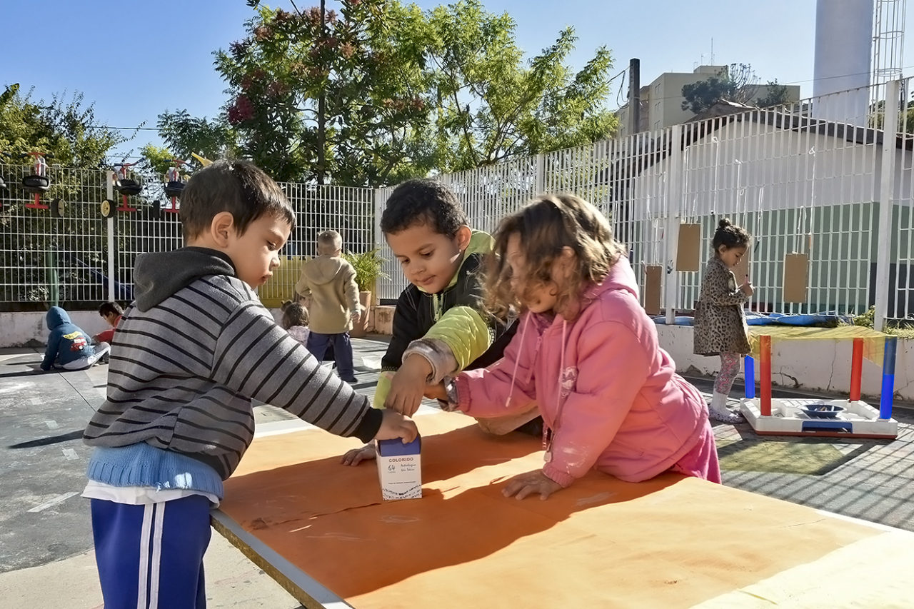 Crianças com agasalhos brincando apoiadas em mesa, em espaço aberto, com céu, árvore e outras crianças ao fundo também brincando