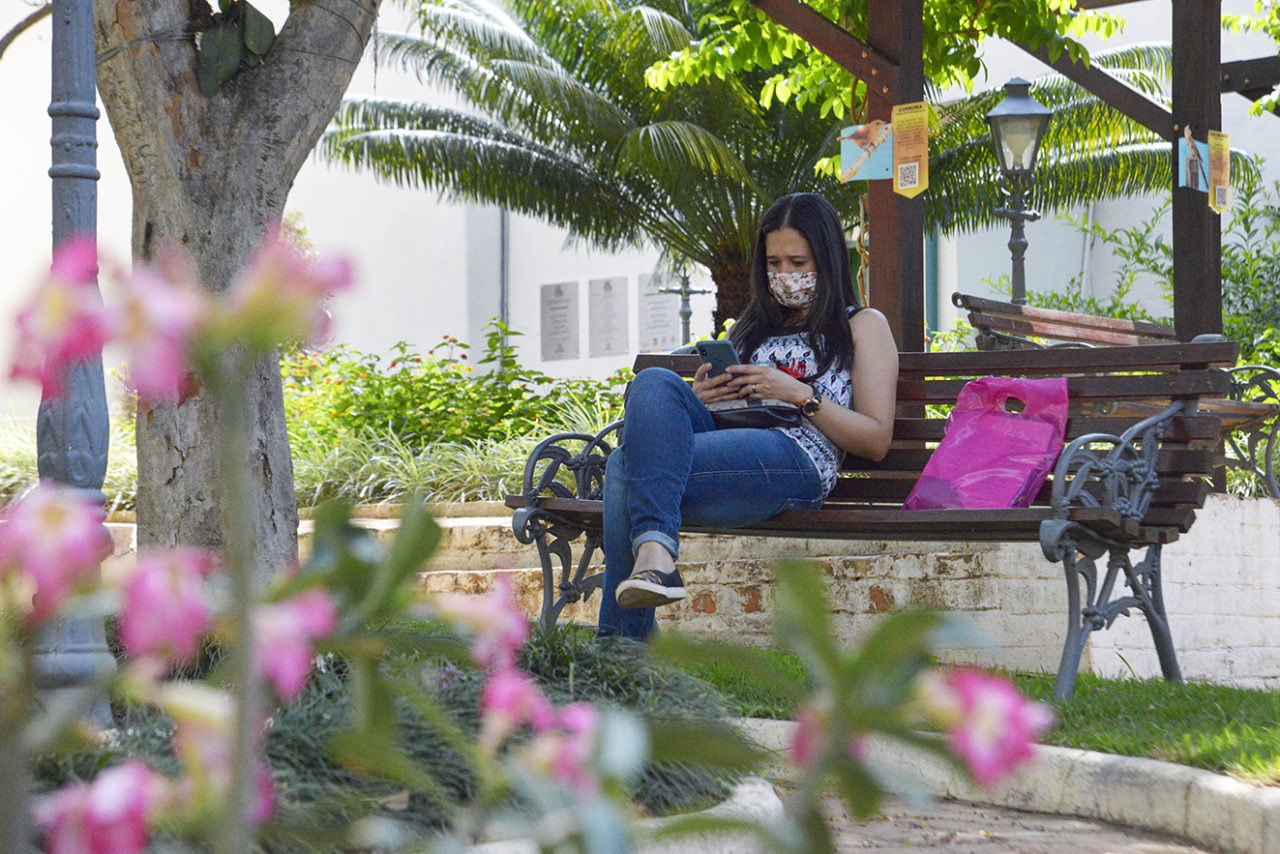 Espaço ao aberto com árvores e flores, com mulher sentada em banco de madeira de praça, usando máscara e mexendo em celular, com sacola ao lado.