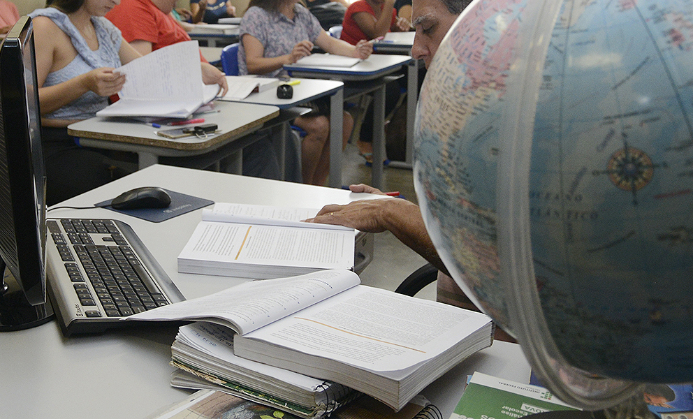 imagem mostra globo em cima da mesa, onde estão livros e cadernos abertos, computador. No fundo há pessoas em carteiras escolares.