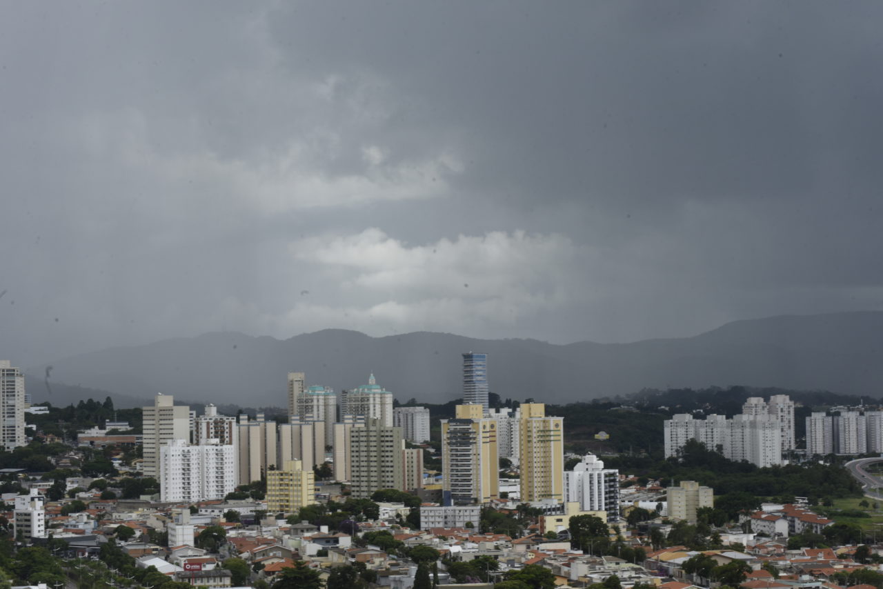 Panorama de cidade com prédios, casas, árvores, e colinas ao fundo, com céu cinzento e nublado, com aspecto de chuva
