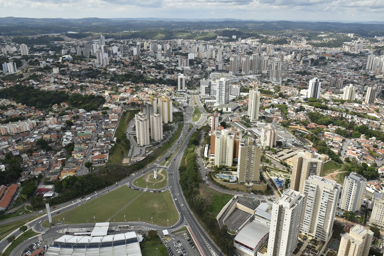 Imagem aérea da cidade mostra avenida com prédios e casas no entorno. 