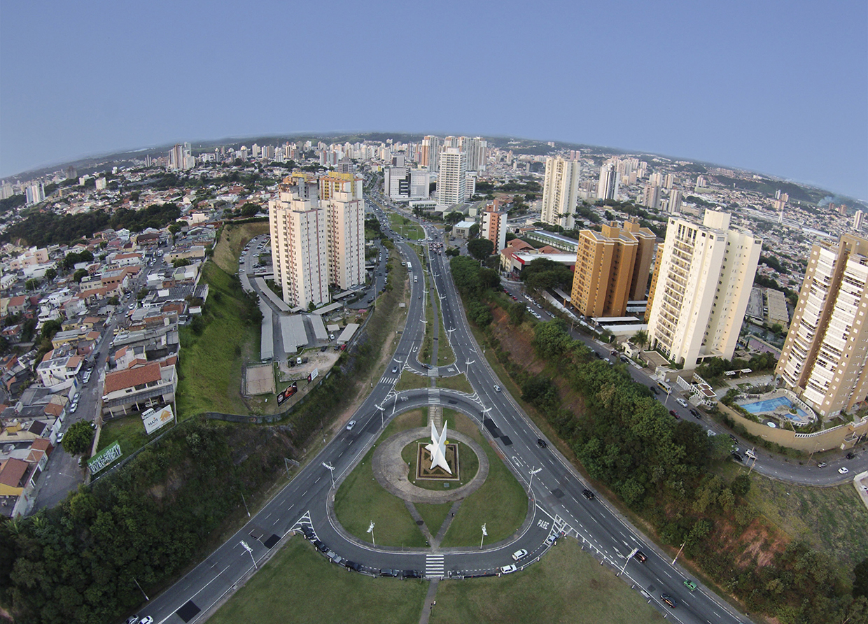 Imagem aérea da cidade, mostra avenidas, rotatória com escultura ao meio, prédios e imóveis no entorno