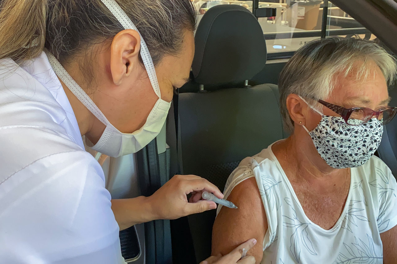 imagem mostra mulher com jaleco branco e máscara aplicando a vacina em idoso que está usando máscara.