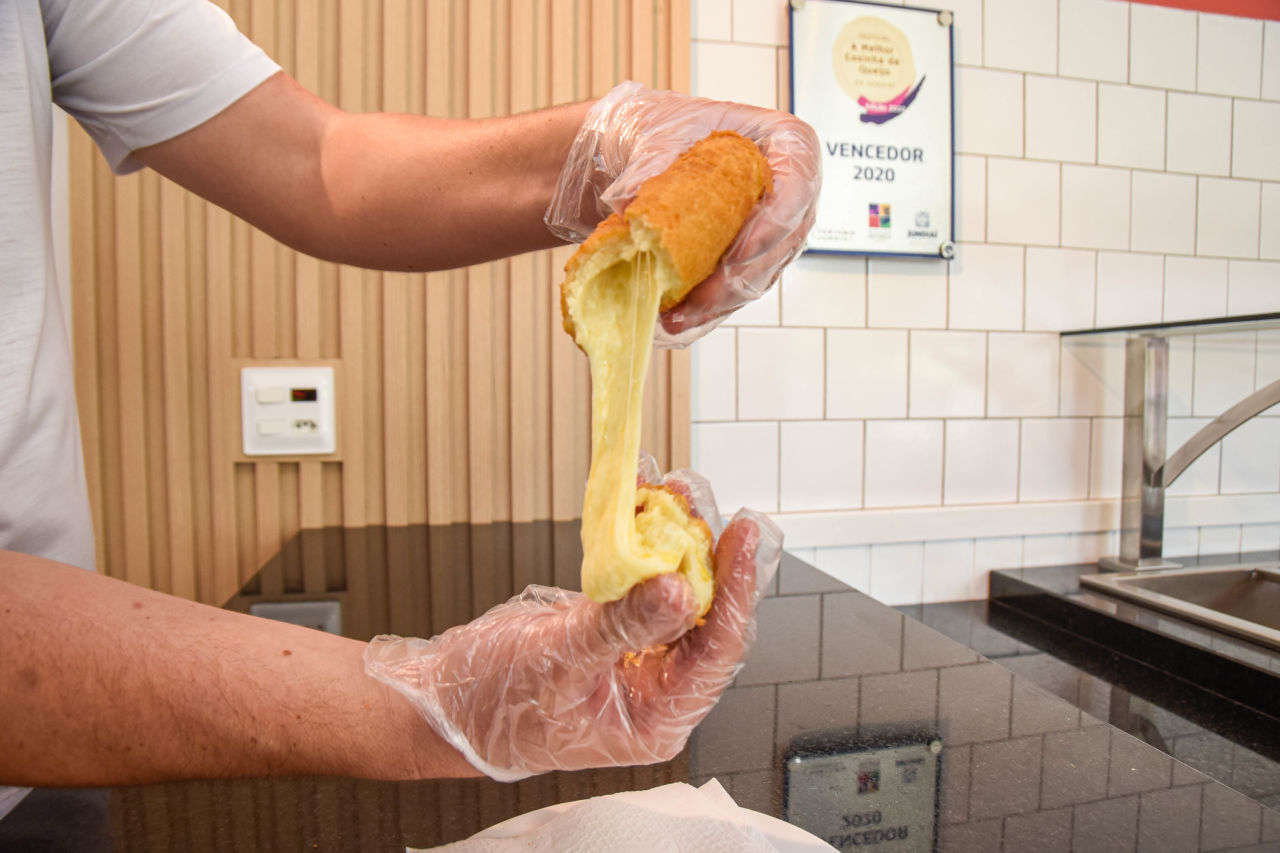 Imagem mostra homem segurando coxinha de queijo nas mãos. O salgado está dividido em dois, sendo cada pedaço em uma mão e o queijo está derretido. 