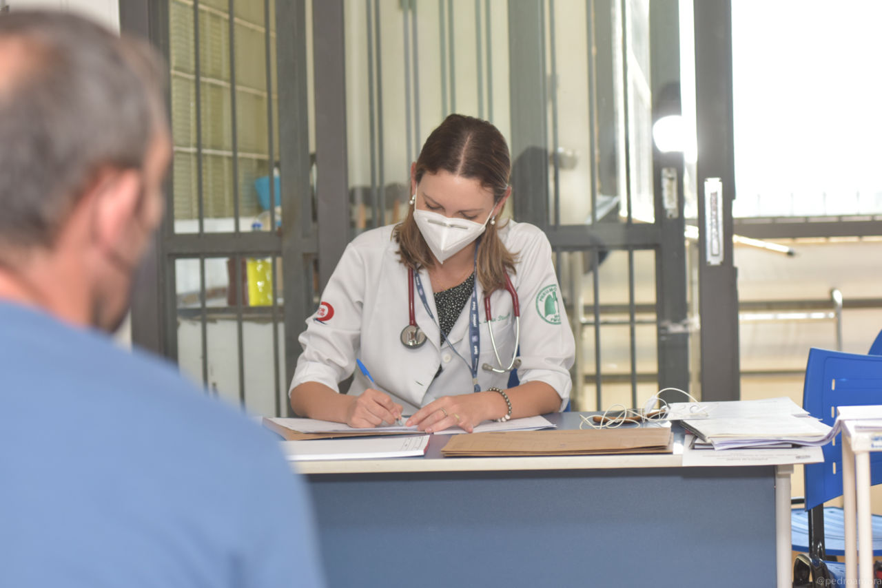 Imagem mostra médica, com estetoscópio no pescoço, jaleco branco, escrevendo em prontuário, enquanto faz atendimento de paciente, que está de costas, com camiseta azul.