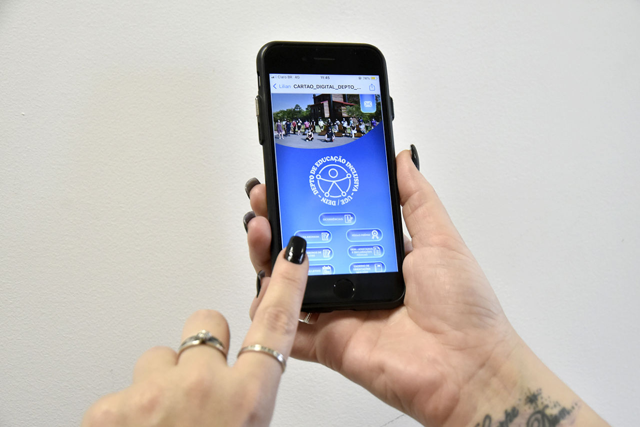 imagem mostra mulher, com as unhas pintadas de esmalte preto, mexendo em celular. Na tela, uma material com fundo azul e ícones para acesso rápido. 