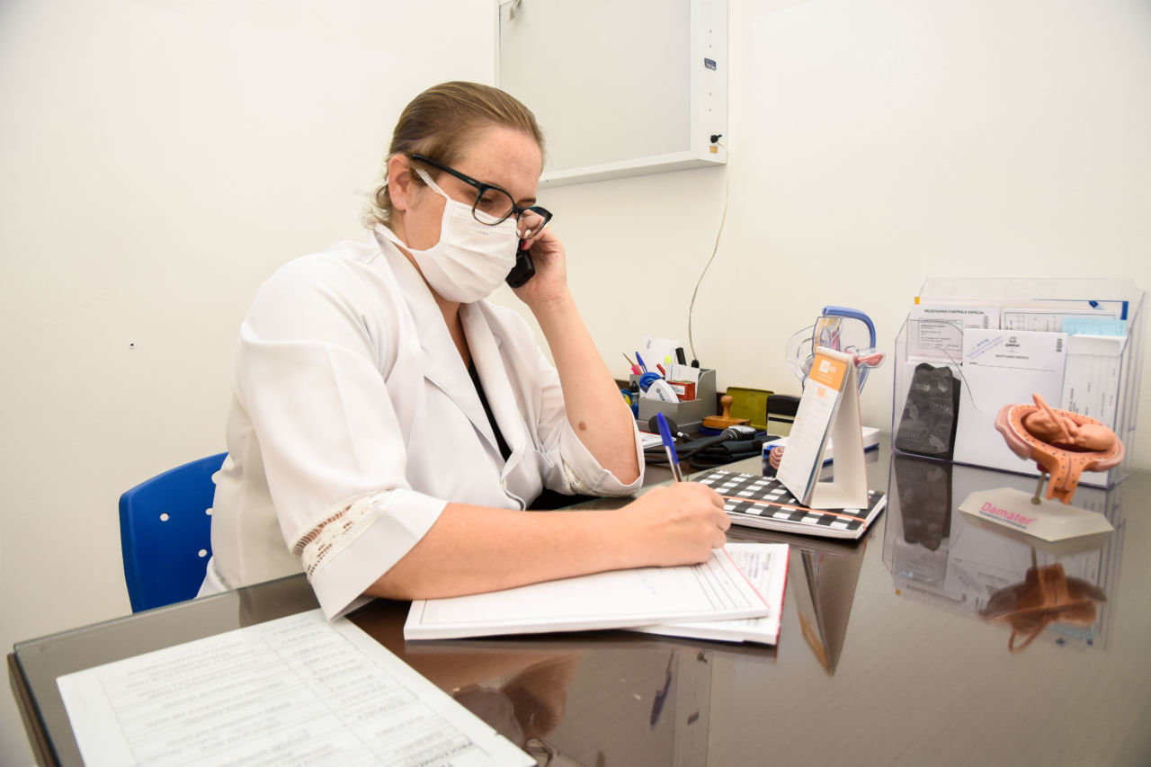 Mulher com jaleco branco sentada em frente à mesa, usando máscara cirúrgica, falando ao telefone e anotando em bloco. 