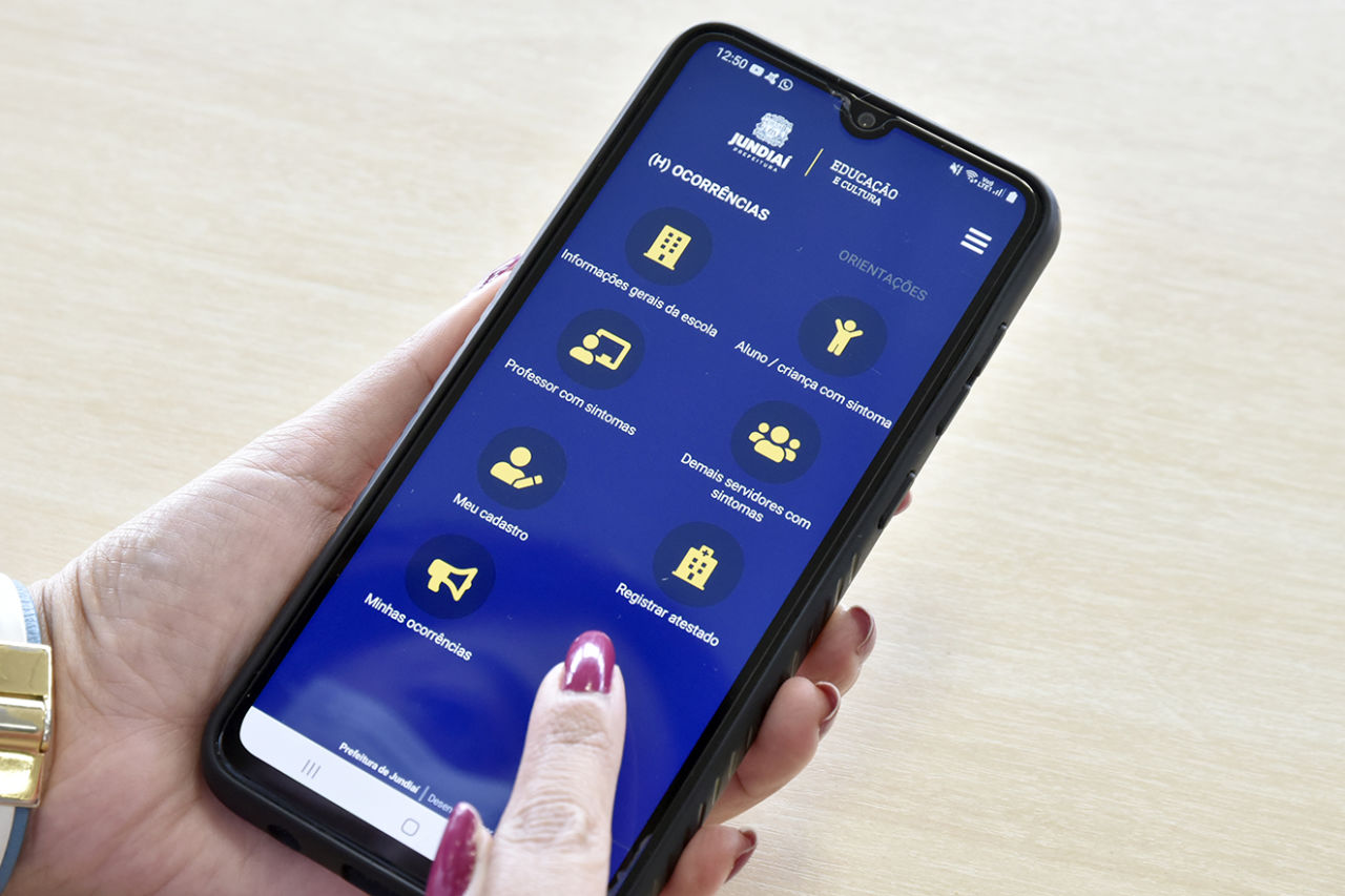 imagem mostra mão de mulher, com celular na mão e na tela do aplicativo da educação, com fundo azul e ícones em amarelo