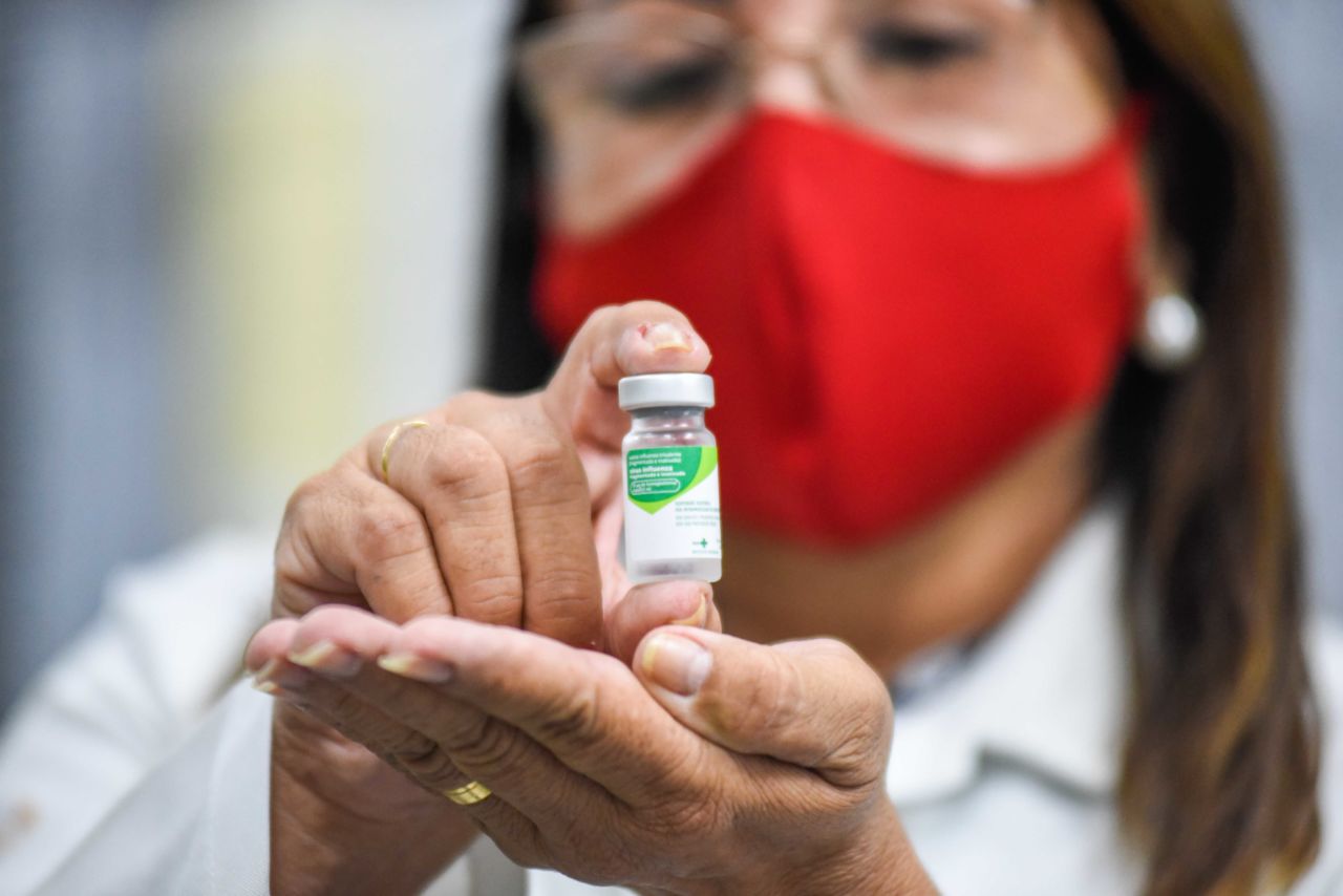 Imagem mostra mulher com máscara vermelha. No detalhe, ela mostra frasco de vacina.
