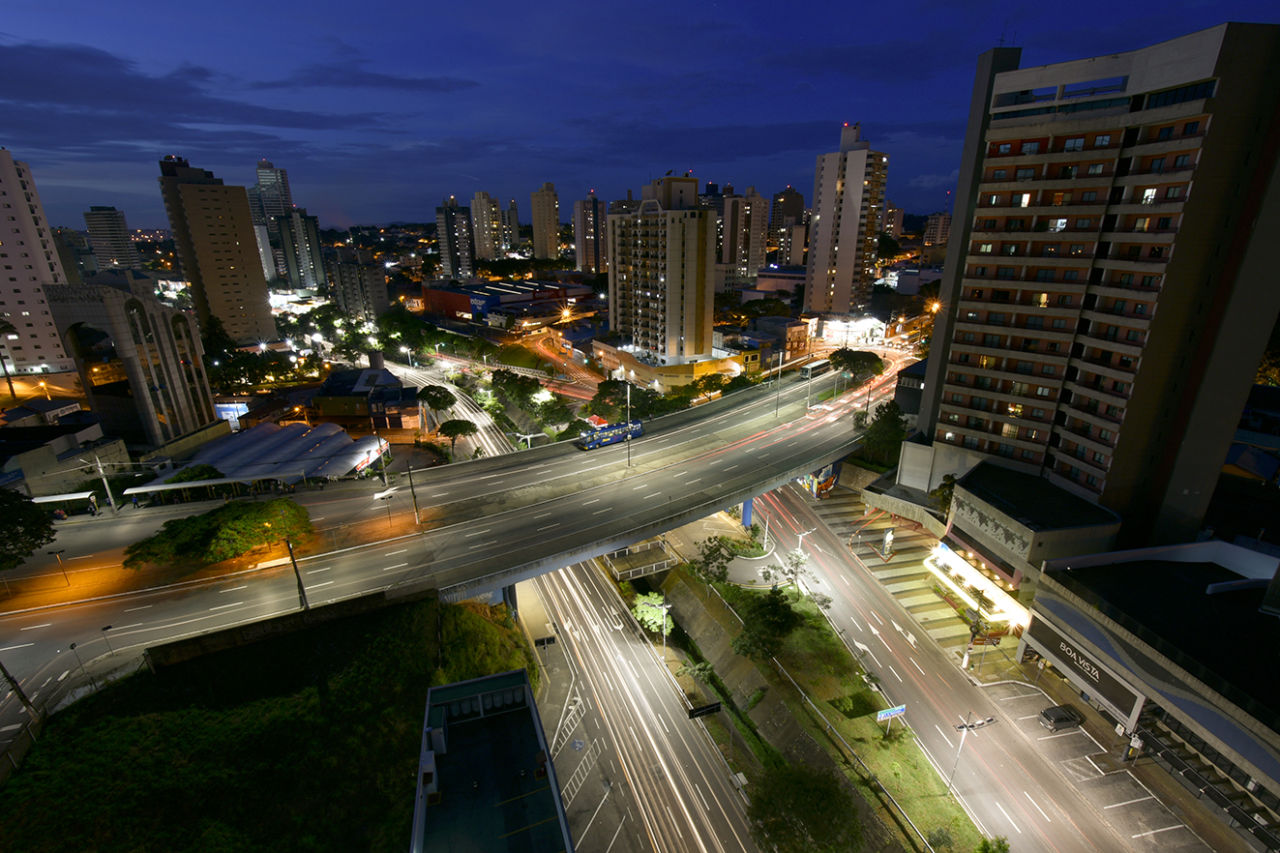 Imagem noturna de cidade iluminada, mostra ponte acima de duas avenidas e um córrego. Nas laterais, diversos prédios com luzes acesas.