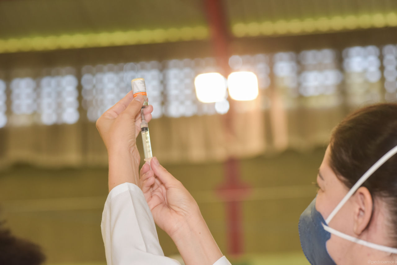Imagem mostra enfermeira com máscara azul, com ampola de vacina e seringa nas mãos. 