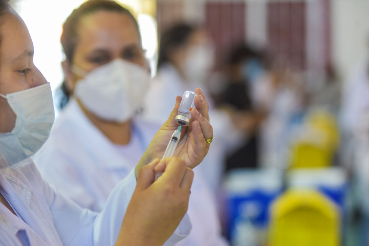 Profissional da Saúde usando máscara  extraindo líquido de ampola com seringa, com fila de outras pessoas ao fundo, desfocado