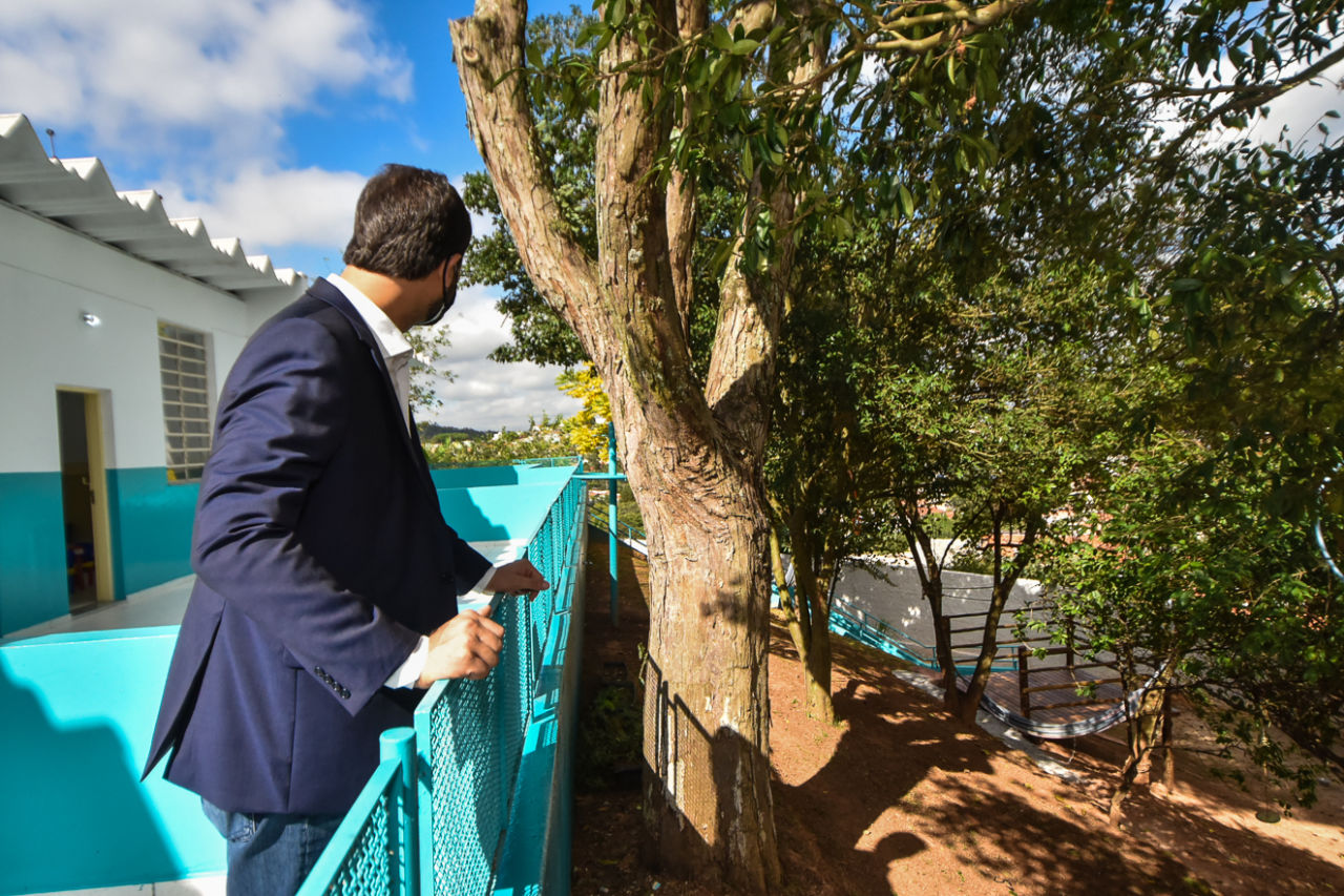 Imagem mostra o prefeito apoiado em grade azul observando espaço externo de escola com deck de madeira e mirante sob árvores