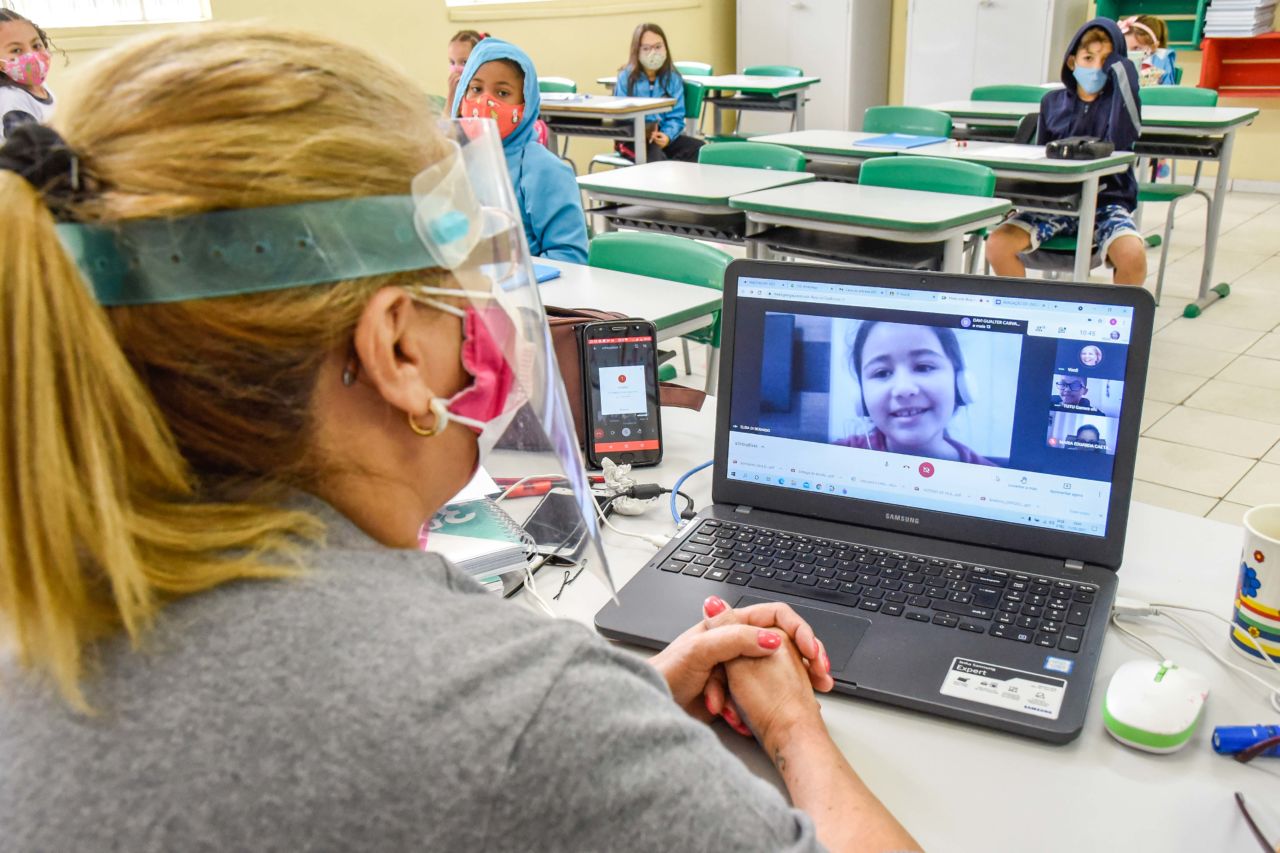 Imagem mostra professora em frente a computador, com alunos em carteiras ao fundo. Na tela, imagem de aluna que conversa com a professora. 