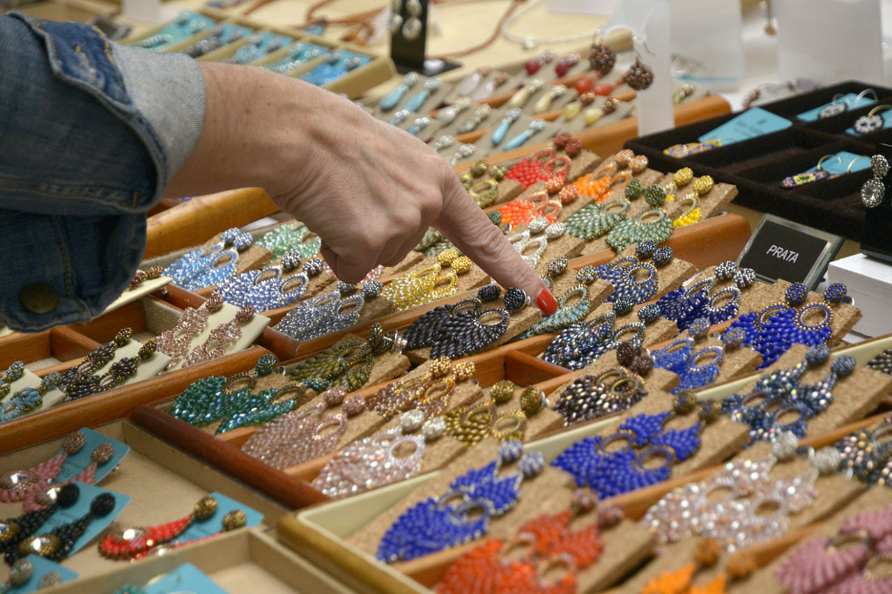 Imagem mostra banca com exposição de bijuterias (brincos coloridos) e mão de molhar, com esmalte laranja apontando para um dos produtos