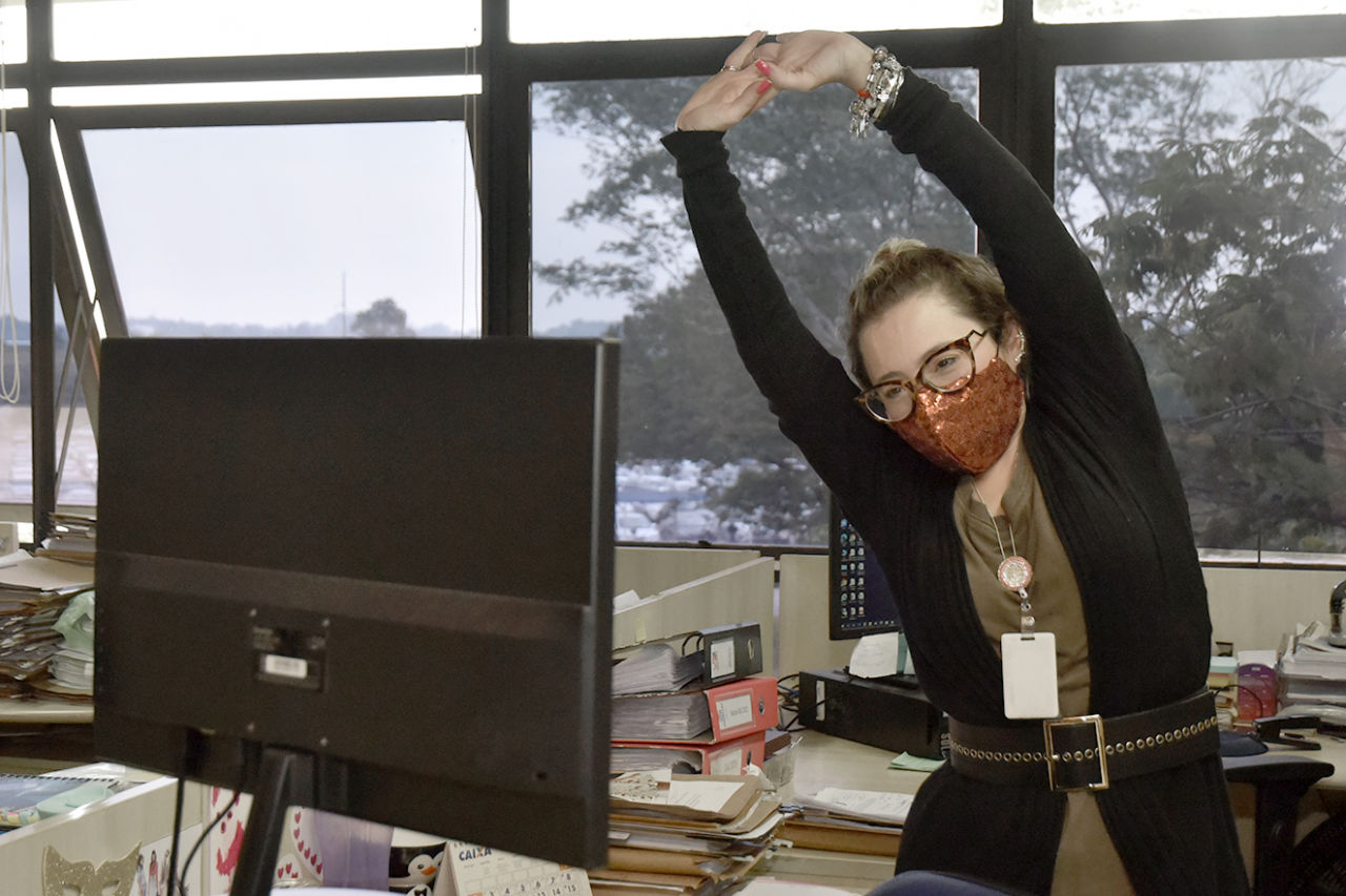 Em ambiente de escritório, mulher com máscara de brilhos e óculos faz exercício levantando os braços em posição de espreguiçar, em pé, olhando para tela de computador.