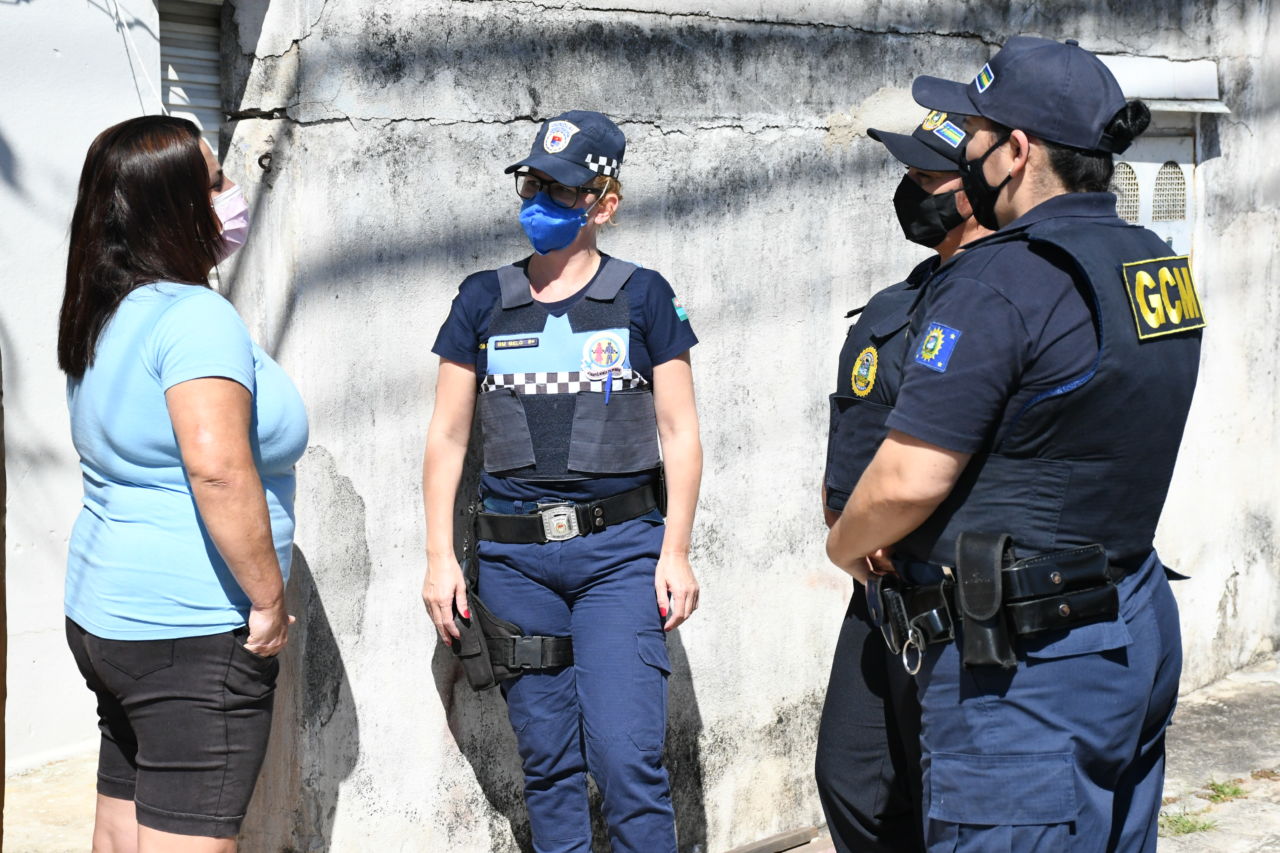 Quatro mulheres de máscaras conversando em pé sobre a calçada em frente a um muro, três delas com roupas de guardas