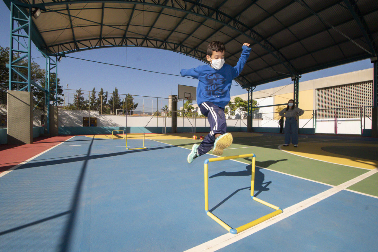 Imagem mostra garoto pulando em pequeno obstáculo próprio para a atividade e idade, em quadra de escola. 