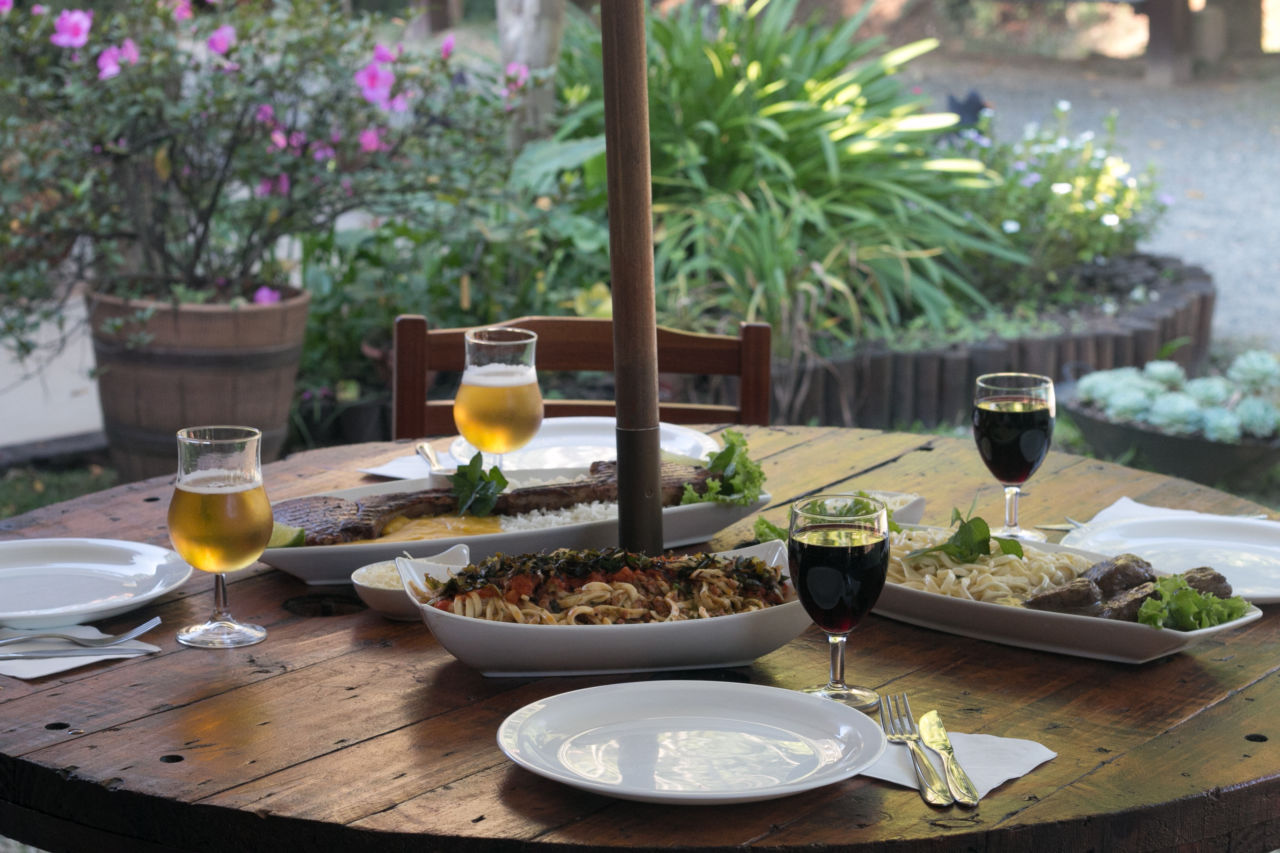 Imagem mostra mesa redonda de madeira, com três pratos com alimentos, macarrão, carnes, dois copos com cerveja e duas taças com vinho tinto.