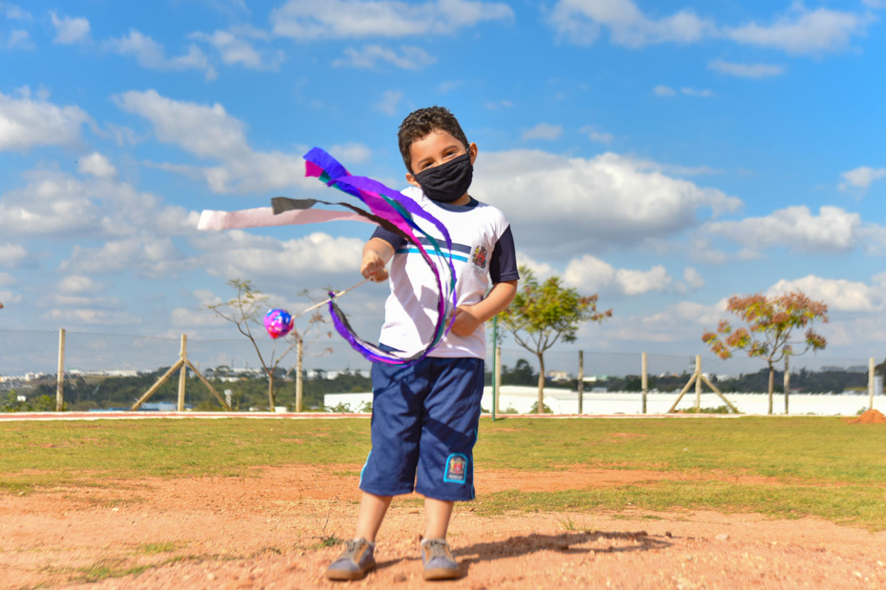 Imagem mostra garoto com uniforme escolar, com brinquedo na mão feito de papéis coloridos e para brincar correndo com o vento