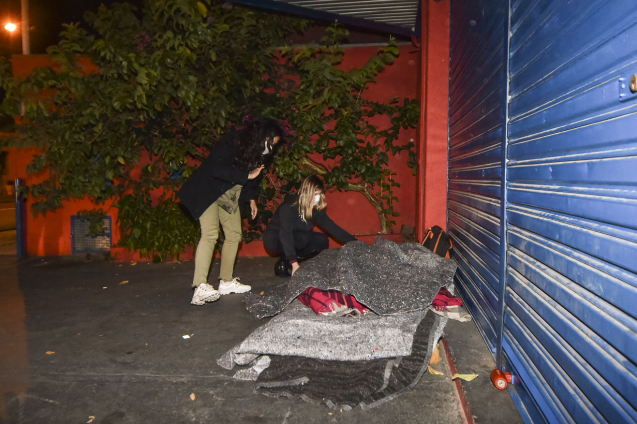 Duas mulheres de máscara estendem cobertor sobre calçada de comércio