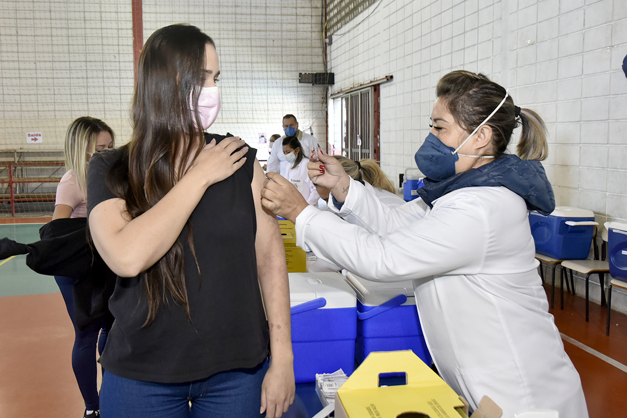 Mulheres usando jaleco e máscara vacina mulher e máscara em pé, dentro de ginásio, com pessoas ao fundo