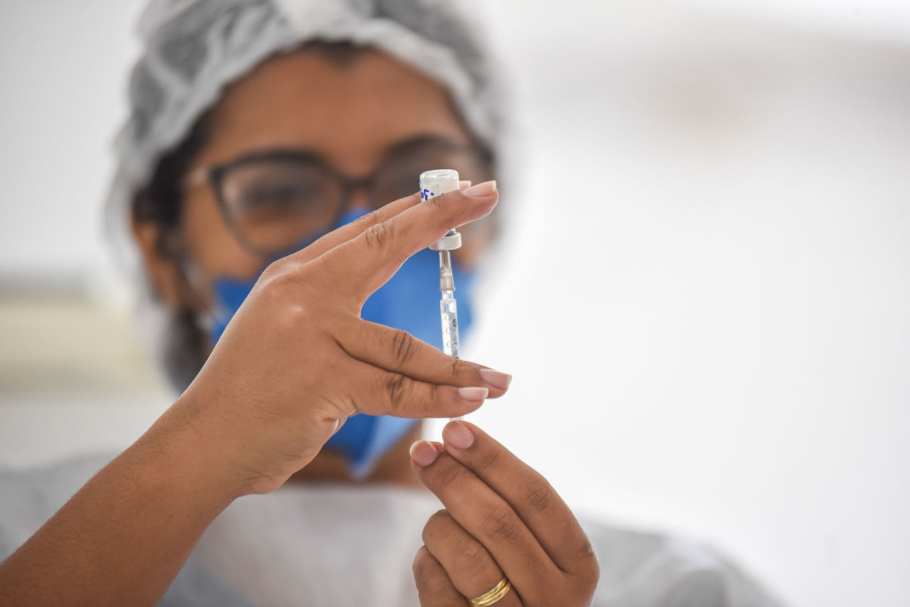 Imagem mostra mulher segurando ampola e tirando vacina com a seringa. 