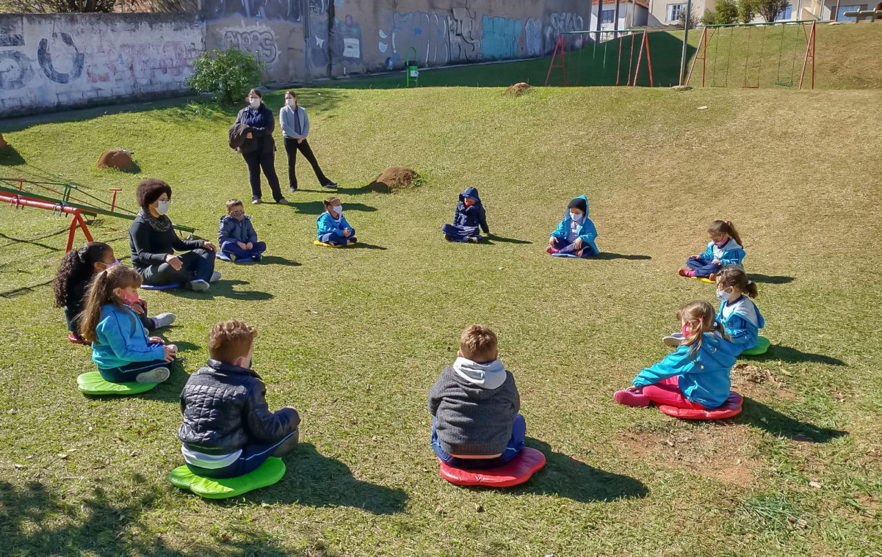 Imagem mostra alunos sentados em círculo, com distanciamento social, em um gramando. Eles participam de uma aula de meditação.
