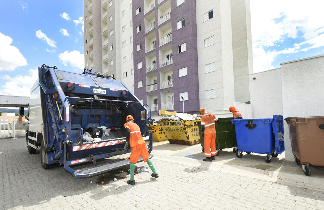 Homens de uniforme no entorno de caminhão, caçambas e contêineres de lixo, com prédio alto ao fundo