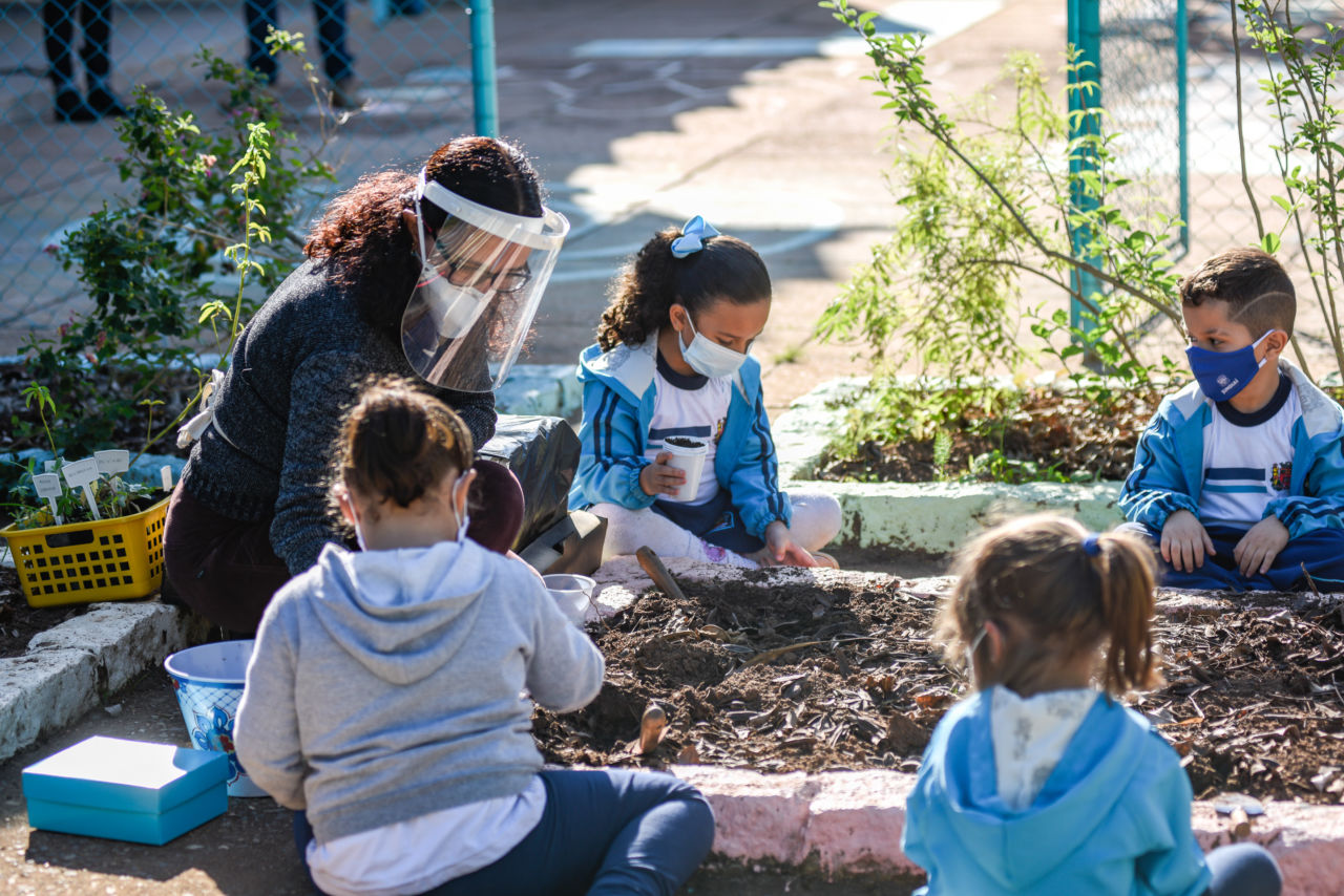 Professora e crianças estão sentadas em volta de um espaço com terra, uma futura horta